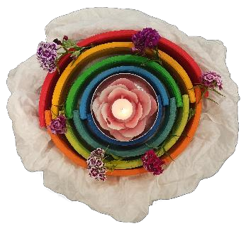 Unsere Kreismitte: Holzringe in Regenbogenfarben, in der Mitte eine Kerze in einer Rosenschale, ringsum Nelkenblüten